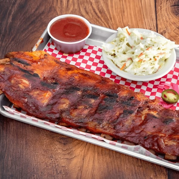 A full rack of BBQ ribs with coleslaw and a side of sauce on a metal tray.