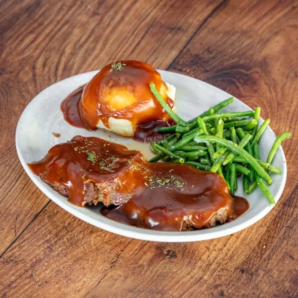 Meatloaf with gravy, mashed potatoes, and green beans on a white plate on a wooden table.