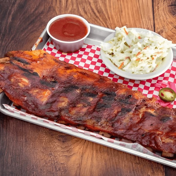 A full rack of BBQ ribs, coleslaw, and dipping sauce on a metal tray with checkered paper.